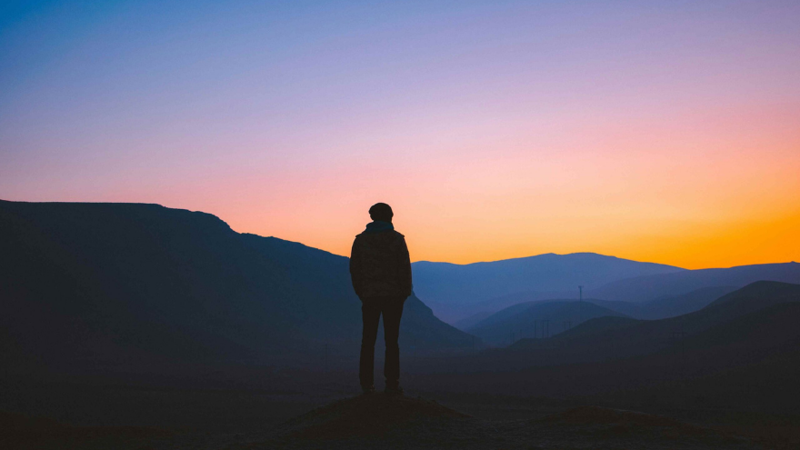 A person standing on a hill looking out at the sun rising behind hills in the distance creating a sky of orange and pinks