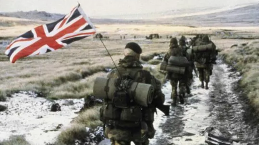Commandos from 40 Commando Anti-Tank Troop march towards Port Stanley in amongst a column of from 45 Commando (the unit to which they were attached). Royal Marine Peter Robinson, carrying the Union Flag attached to the aerial of the radio he is carrying, brings up the rear.