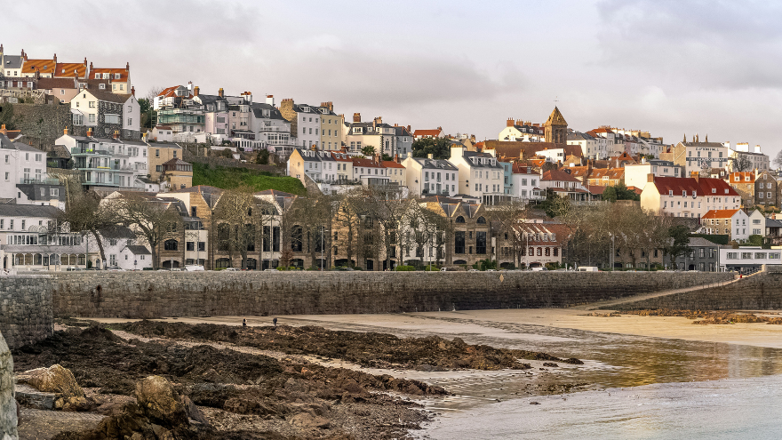 Guernsey coastal townscape with a line of quaint, colourful houses atop a hill, overlooking a rocky shore and calm sea under a cloudy sky.