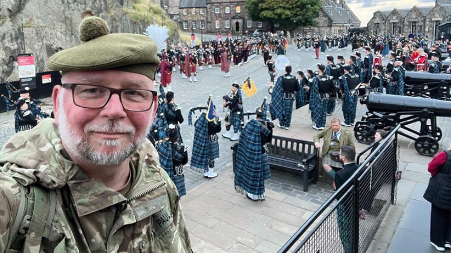 ASR Paul Curd taking a selfie in front of a large group of uniformed bagpipers practicing in a courtyard.